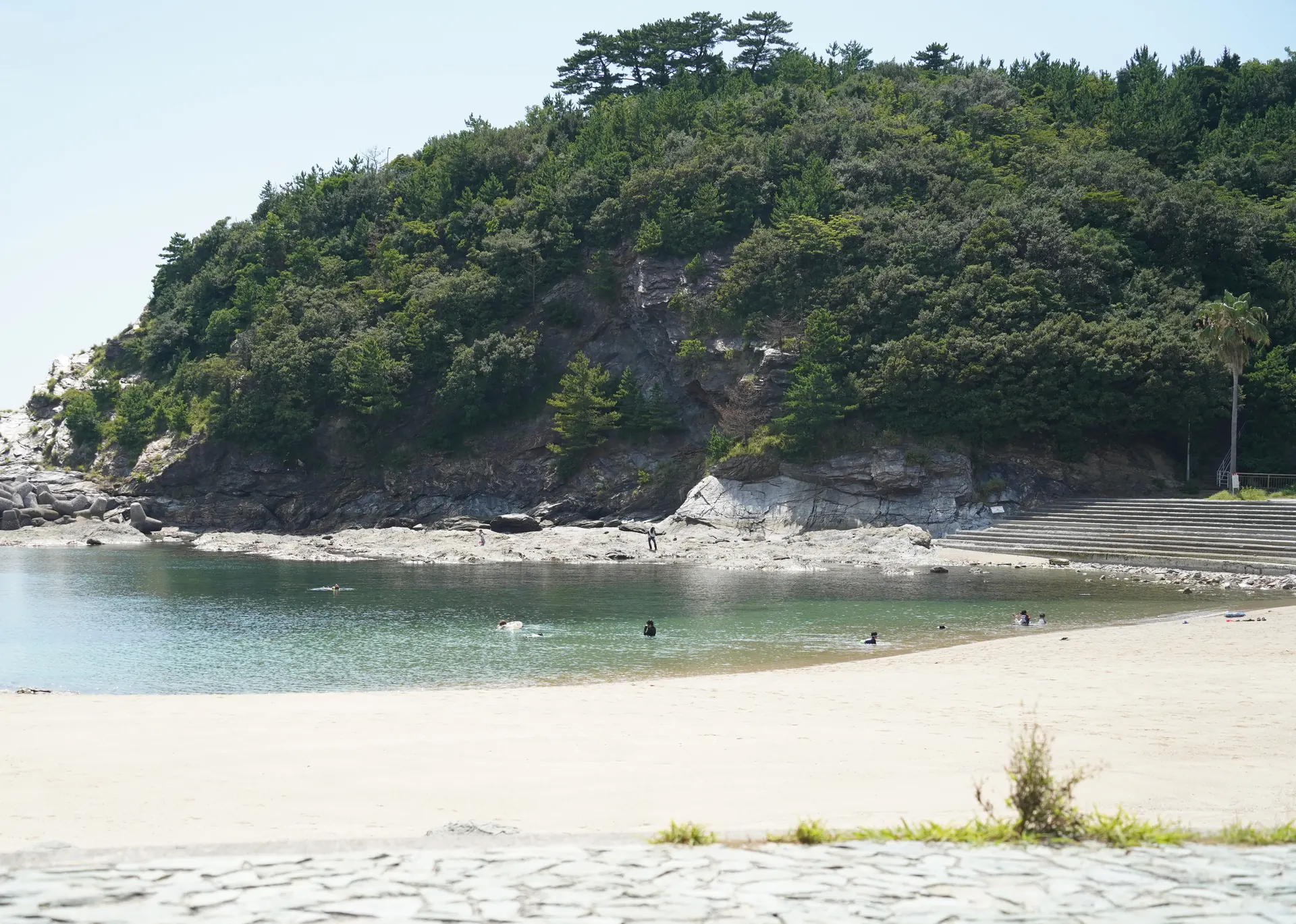 夏の日中の浪早ビーチ。手前に広い砂浜、奥は入り江の穏やかな浅瀬で、その先に松の茂る岩の崖と浜へ下りる階段が見える
